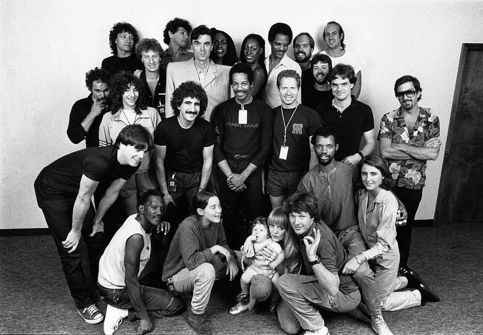 A large group photo in black and white: about twenty-five people in rows, David Byrne in a pale jacket near the centre