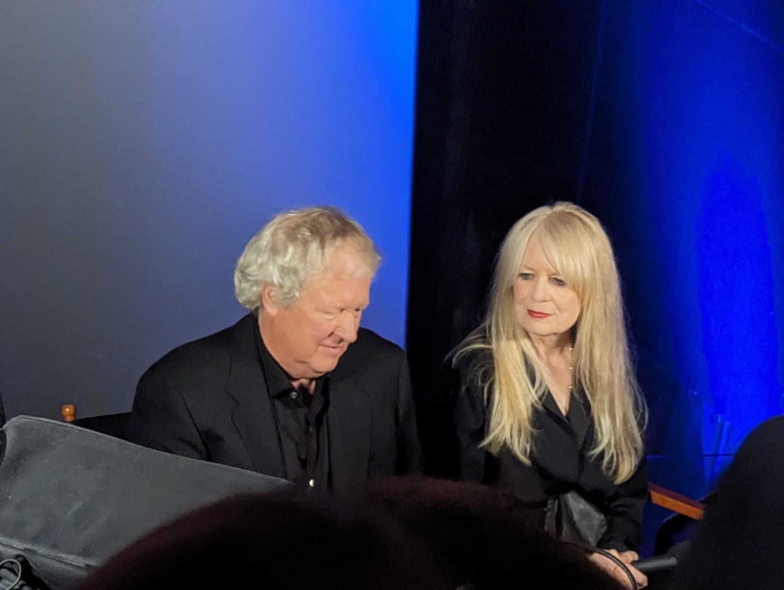 A man with white hair and a woman with long blonde hair seated in a cinema under blue light