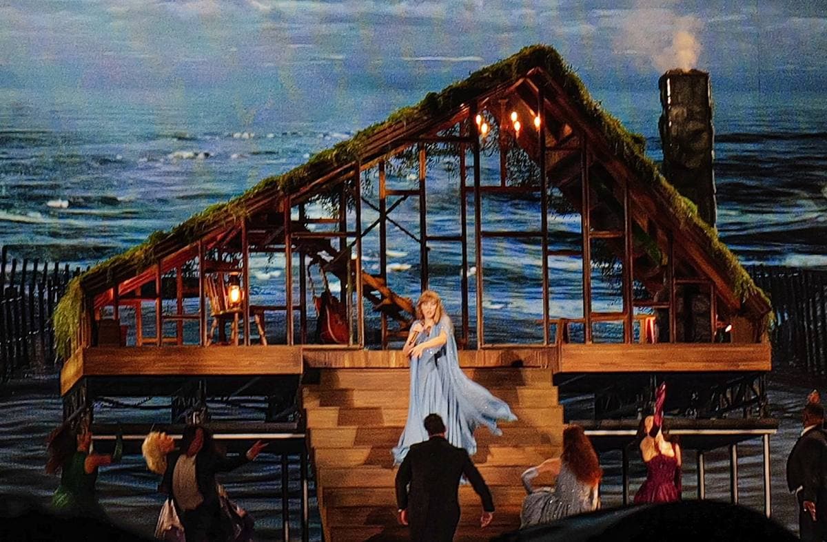 A wooden cabin with a mossy roof on a stadium stage, Taylor Swift in a pale gown on its steps