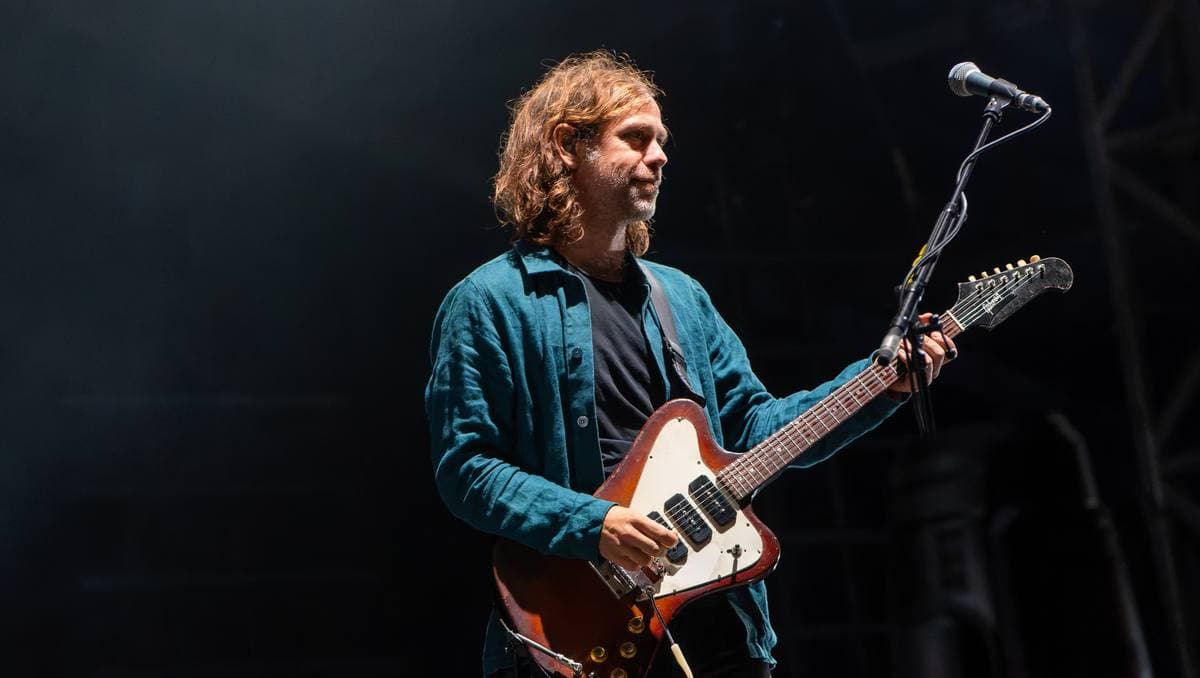 Aaron Dessner with curly hair playing a red guitar against a dark stage