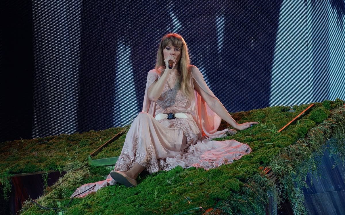 Taylor Swift lying on a moss-covered stage roof in a long pale dress