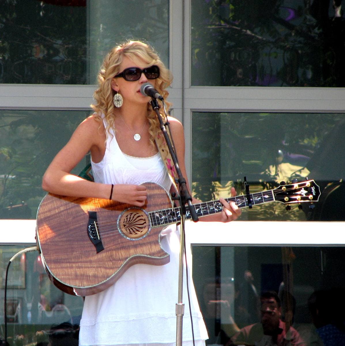 A teenage Taylor Swift in a white dress and sunglasses, singing with an acoustic guitar