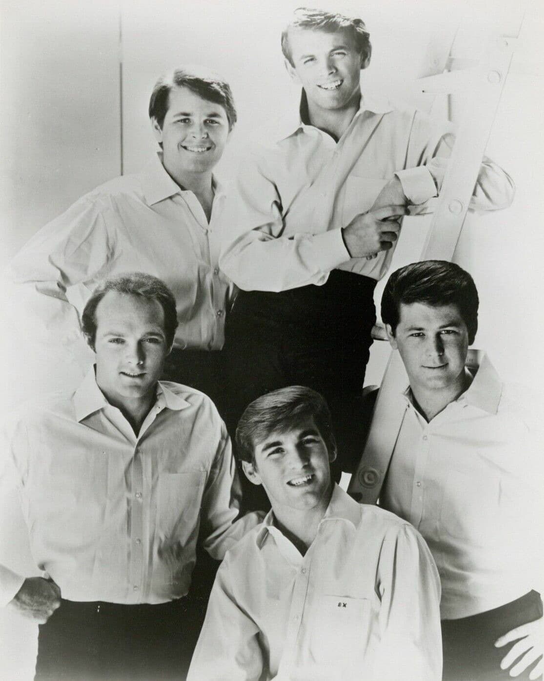 Five young men in pale shirts posed around a ladder in a studio, smiling