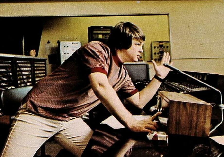 A young man in a striped shirt leans over a mixing desk, one hand raised towards the studio glass