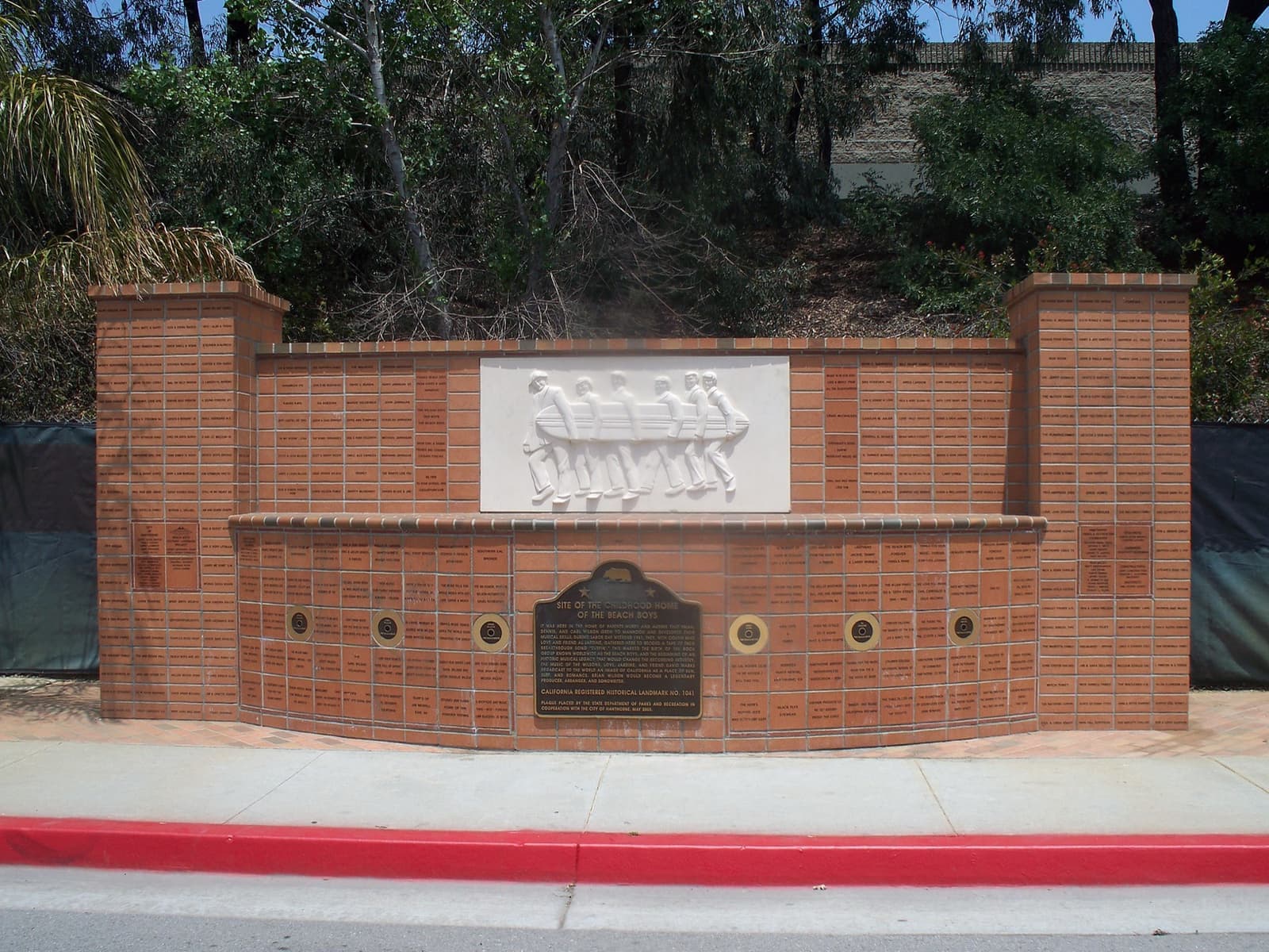 A curved brick wall with a white relief of five surfers carrying a board, a bronze plaque and gold discs set into the brick