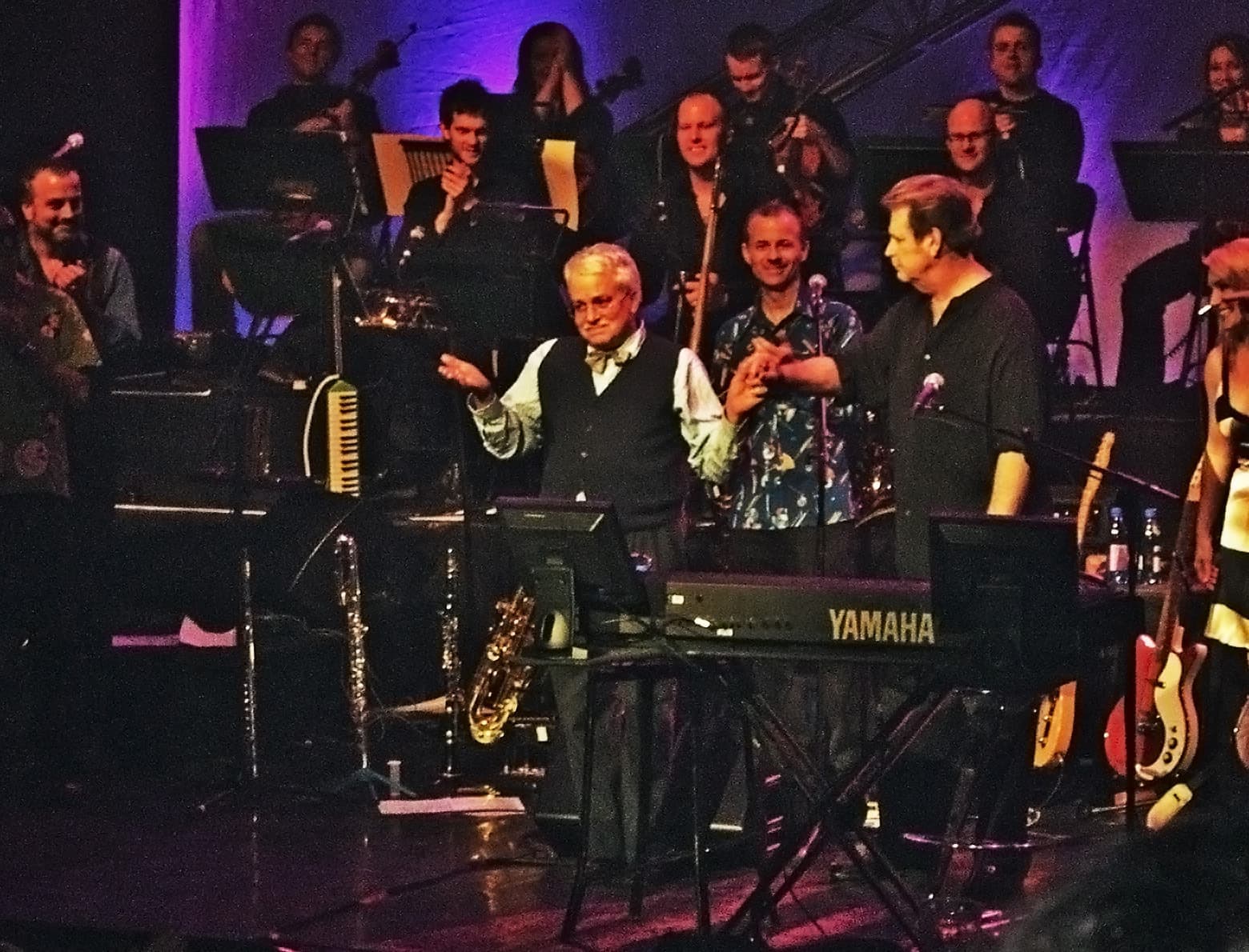 Two men on a stage among musicians and stands, one at a keyboard