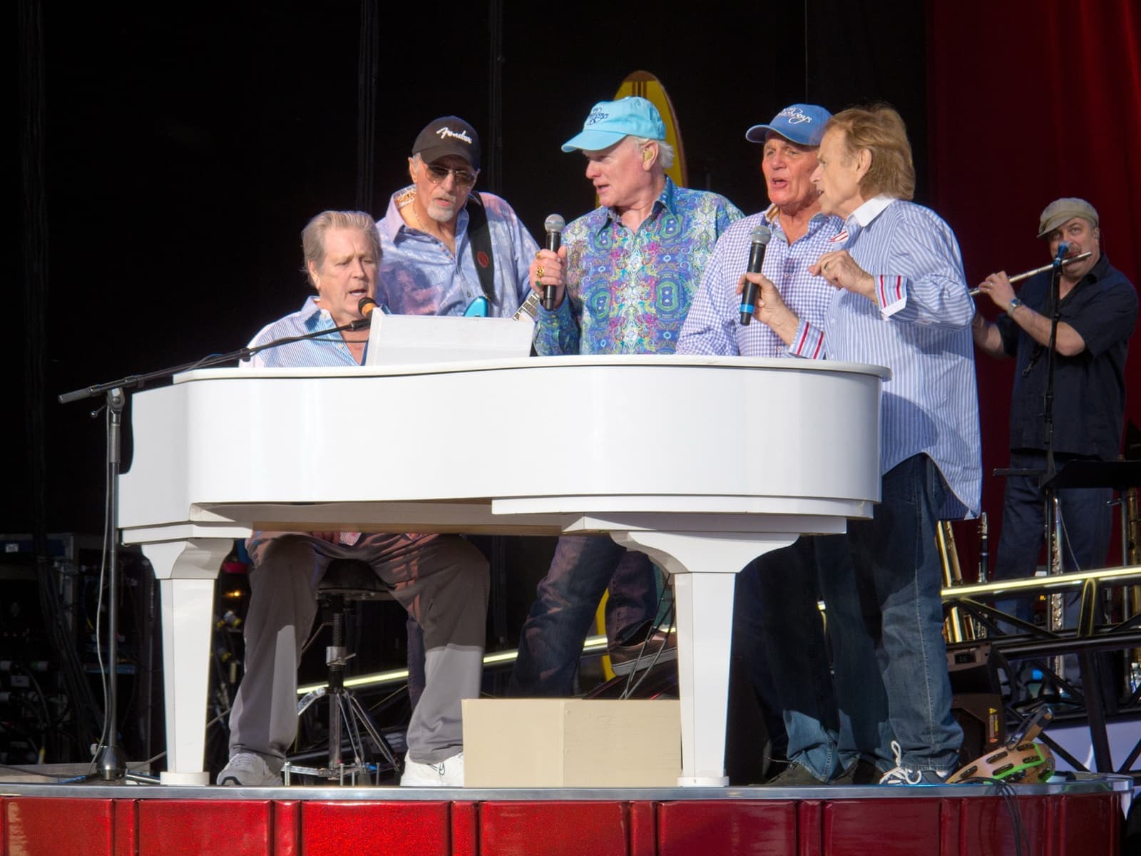 Five older men in caps and shirts singing together around a white grand piano on stage