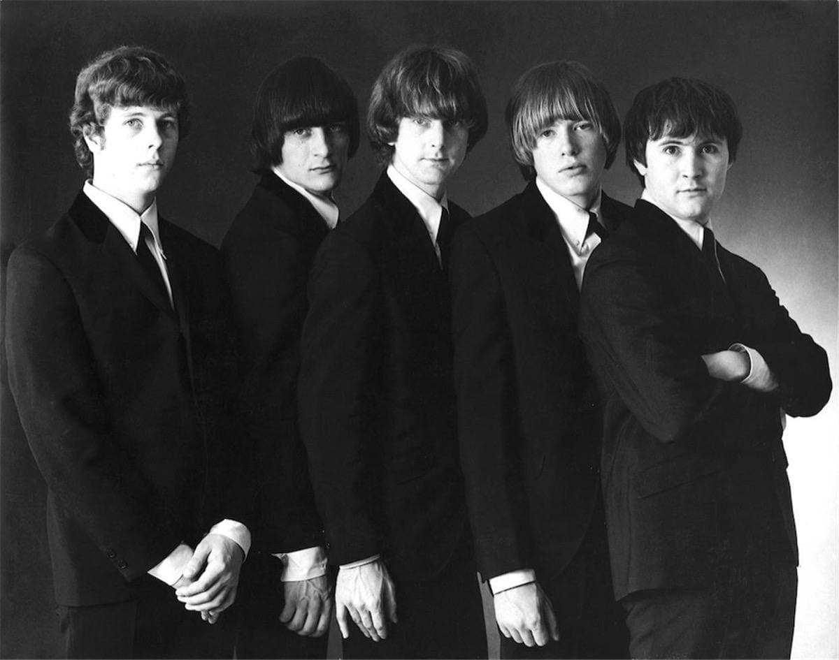 Five young men with long fringes in dark suits, standing in a line against a dark background, black and white