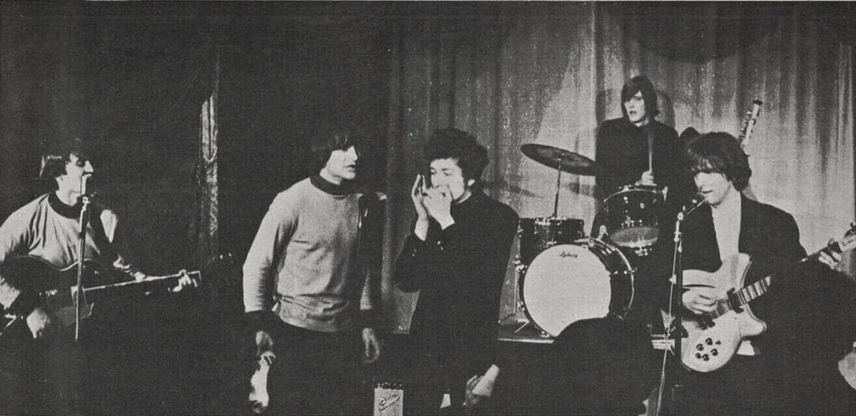 A band on a small stage with a drum kit; a man with curly hair plays harmonica among the guitarists, black and white