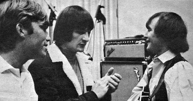 Three young men talking in a recording studio in front of an amplifier, one holding a guitar, black and white
