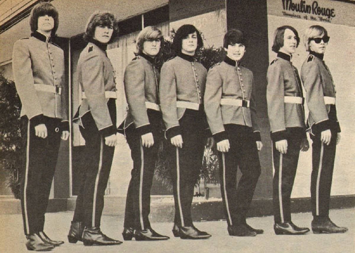 A row of young men in guardsman-style uniforms outside a club with Moulin Rouge on the wall, black and white