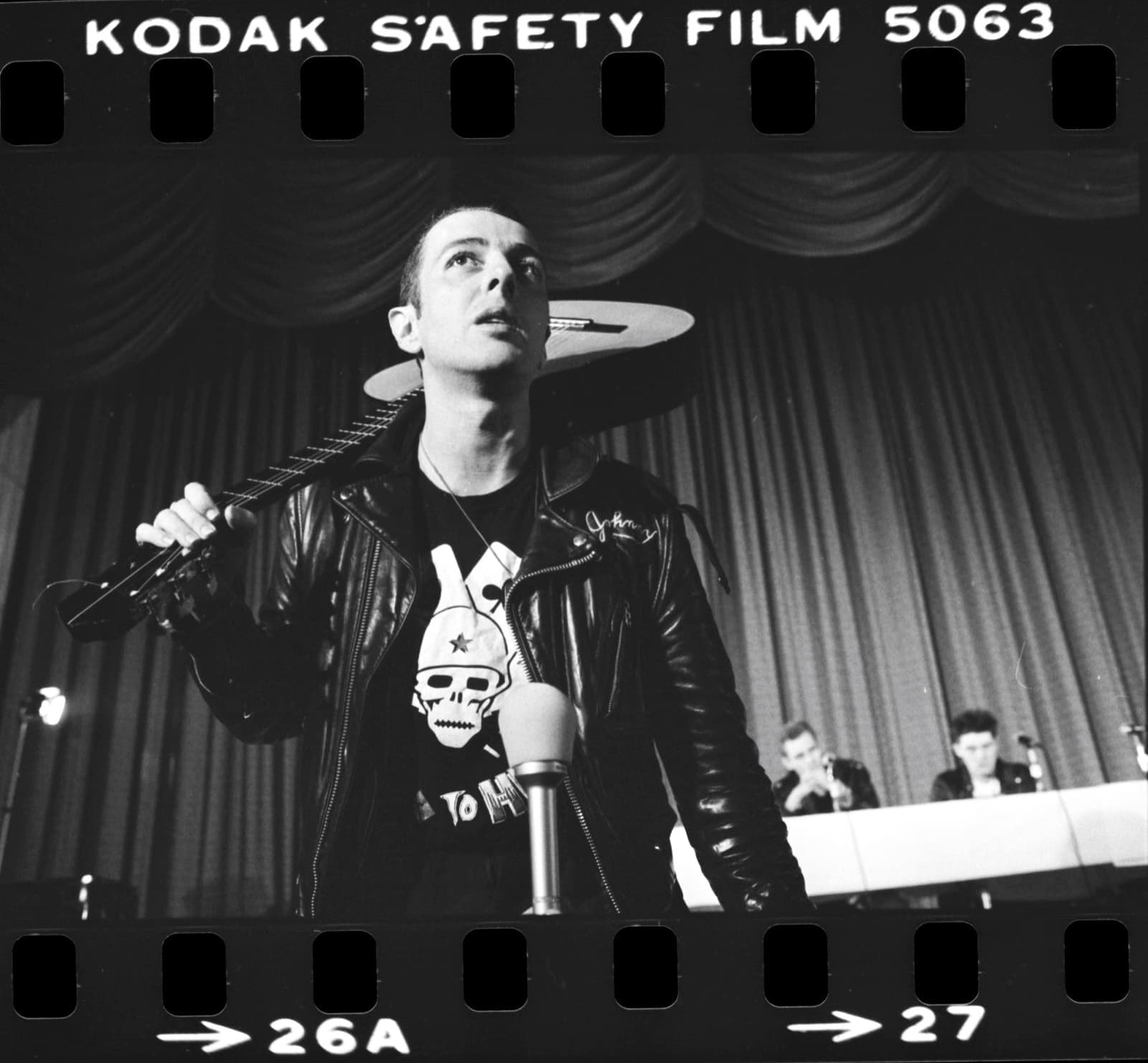 Joe Strummer in a leather jacket holding a microphone stand on a stage, on a contact-sheet frame