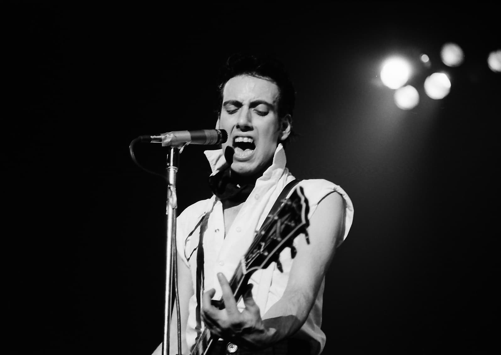 Mick Jones singing and playing a guitar under stage lights