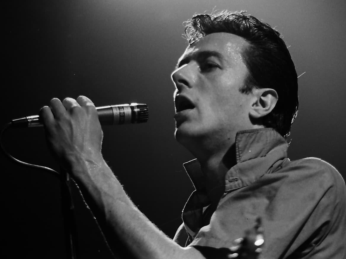 Joe Strummer singing into a microphone under a stage light, eyes half closed, in black and white