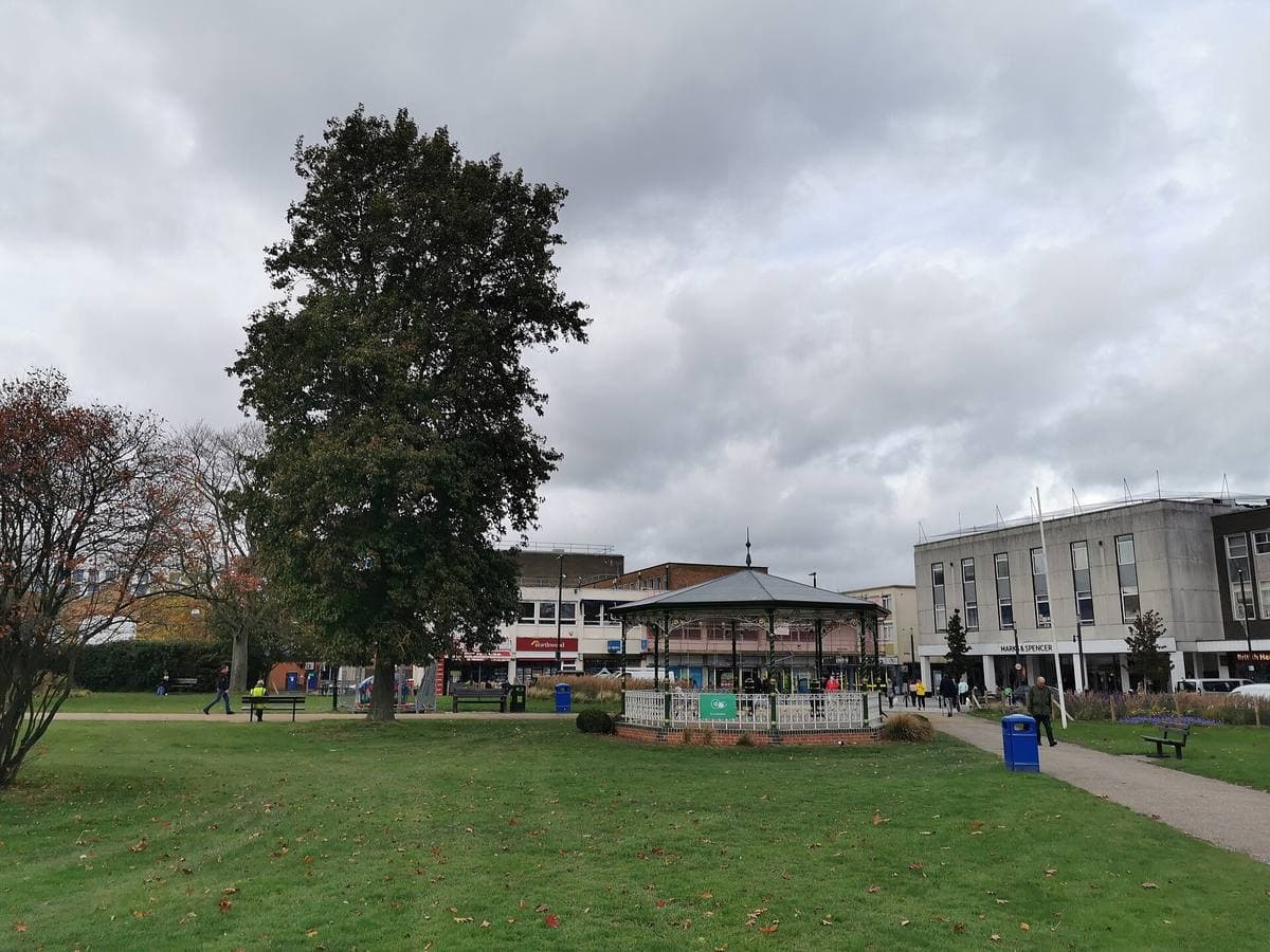 A small octagonal bandstand on a green lawn, with a tall tree beside it and town-centre buildings behind