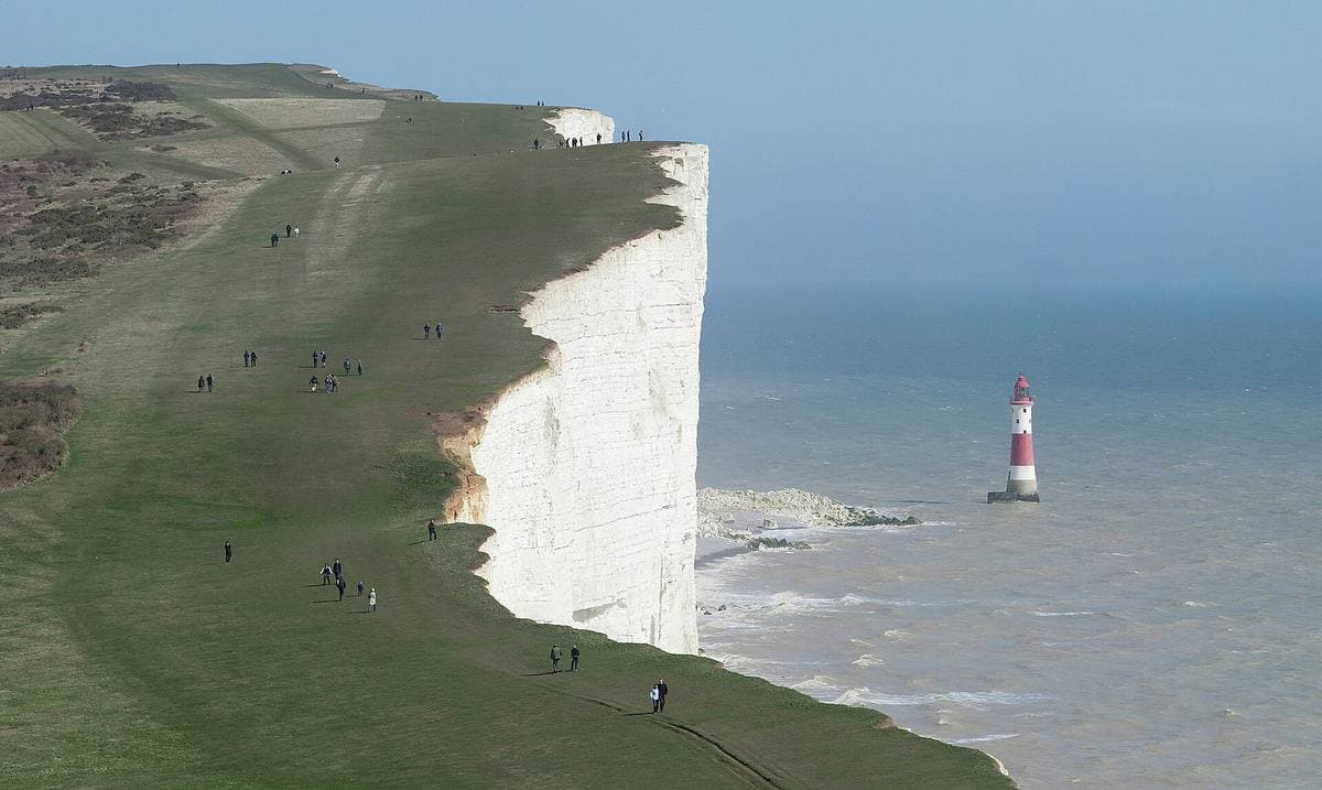 A sheer white chalk cliff dropping into a calm sea, a red-and-white lighthouse below and walkers on the green clifftop