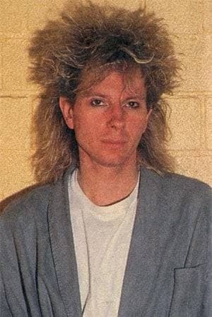 Boris Williams in 1986, a big shock of bleached spiky hair, a grey jacket over a white T-shirt, against a pale brick wall