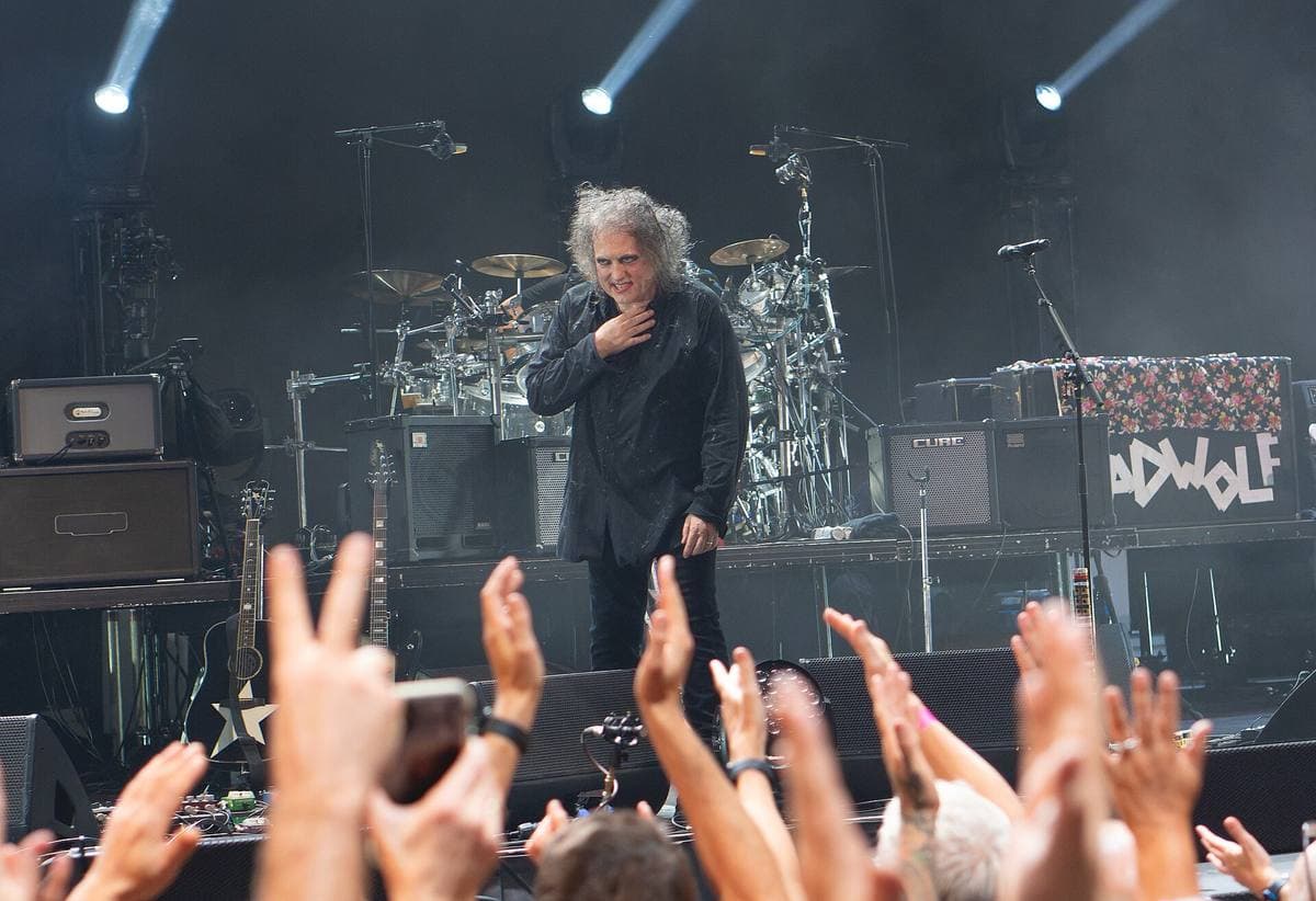 A crowd’s raised hands and phones in front of a lit stage where Robert Smith stands with a hand at his chest