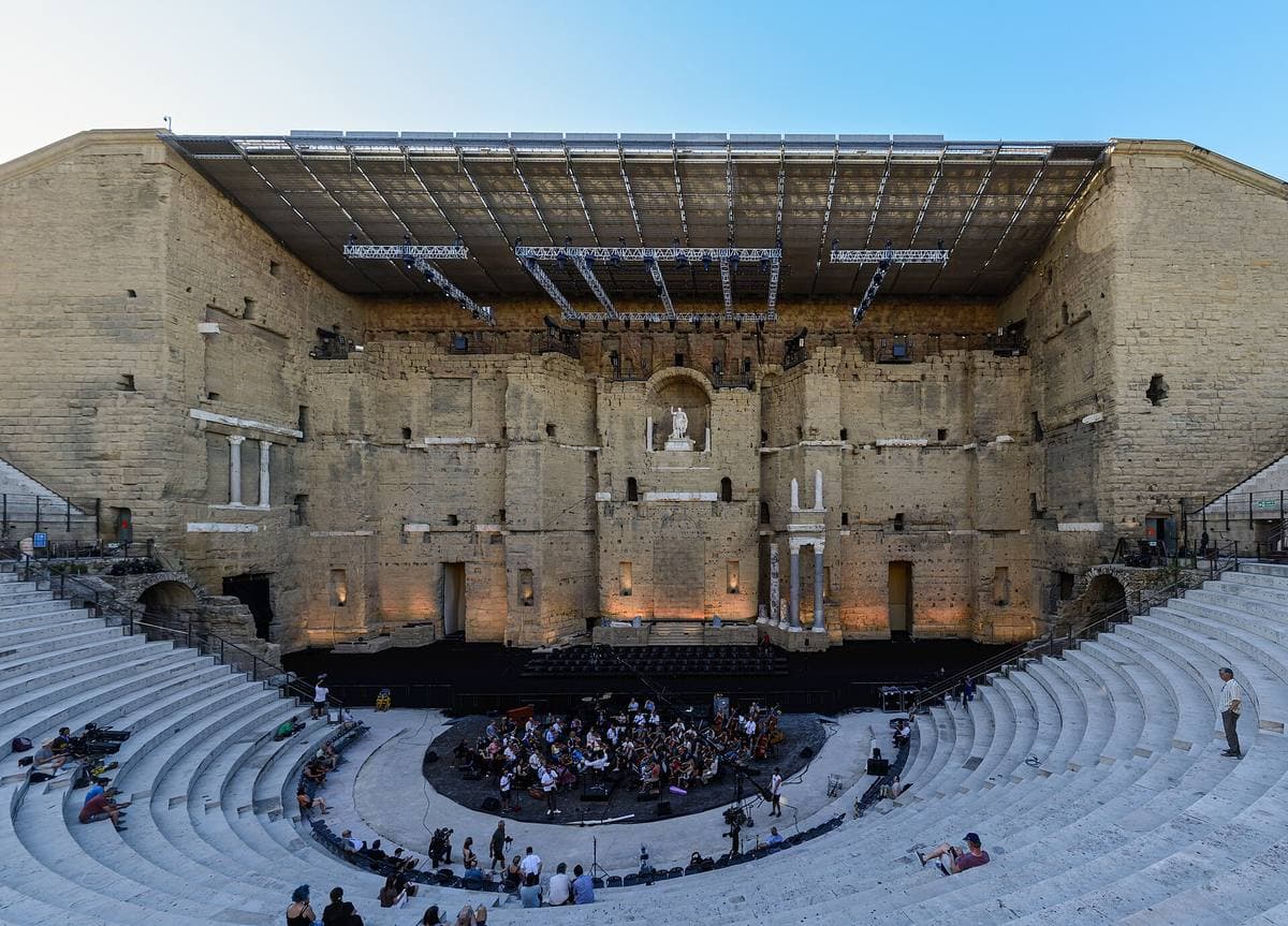A Roman theatre seen from high in its stone seats, the huge sandstone stage wall lit behind an orchestra setting up