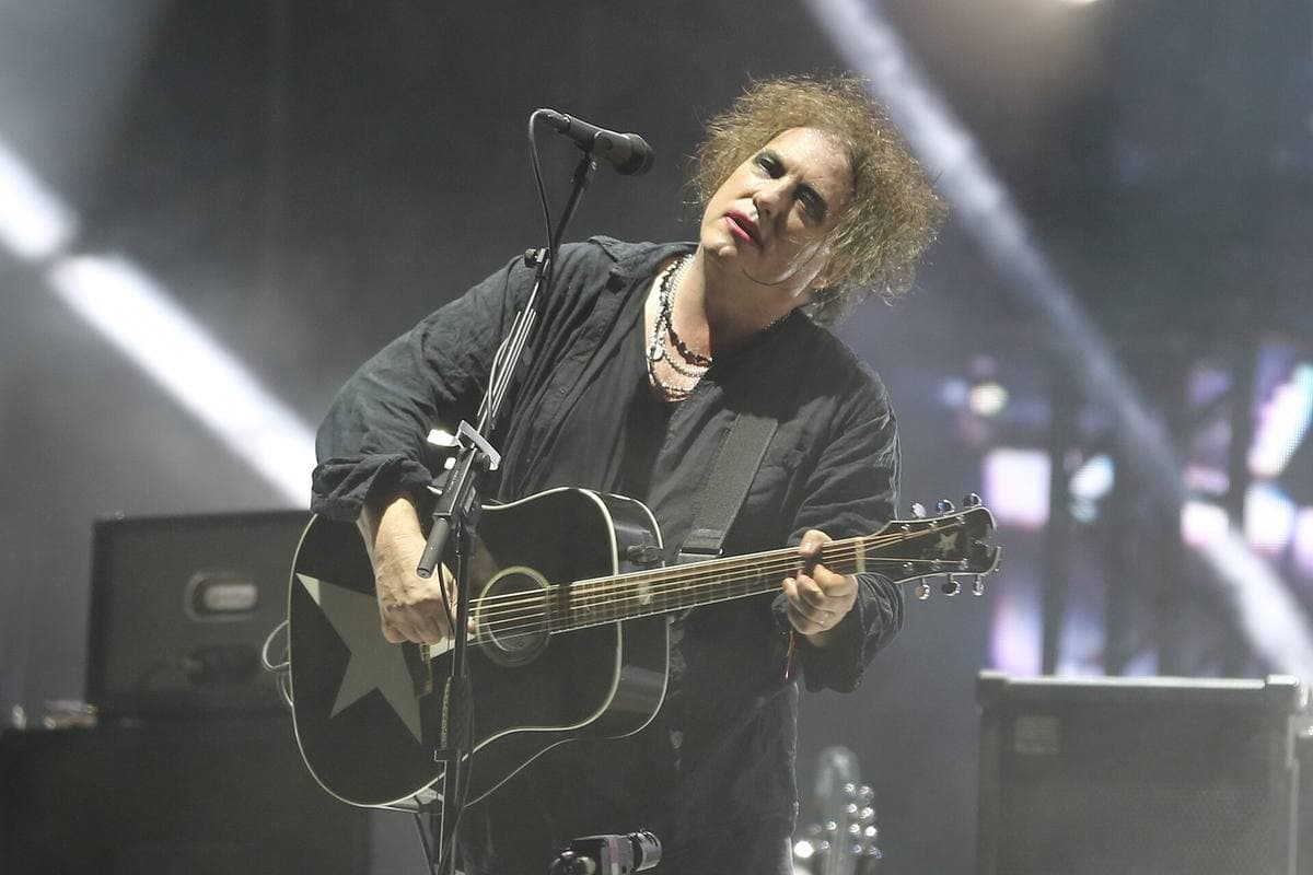 Robert Smith in 2019 singing into a microphone with a black acoustic guitar, his hair a grey-black cloud, red lipstick, white stage lights behind