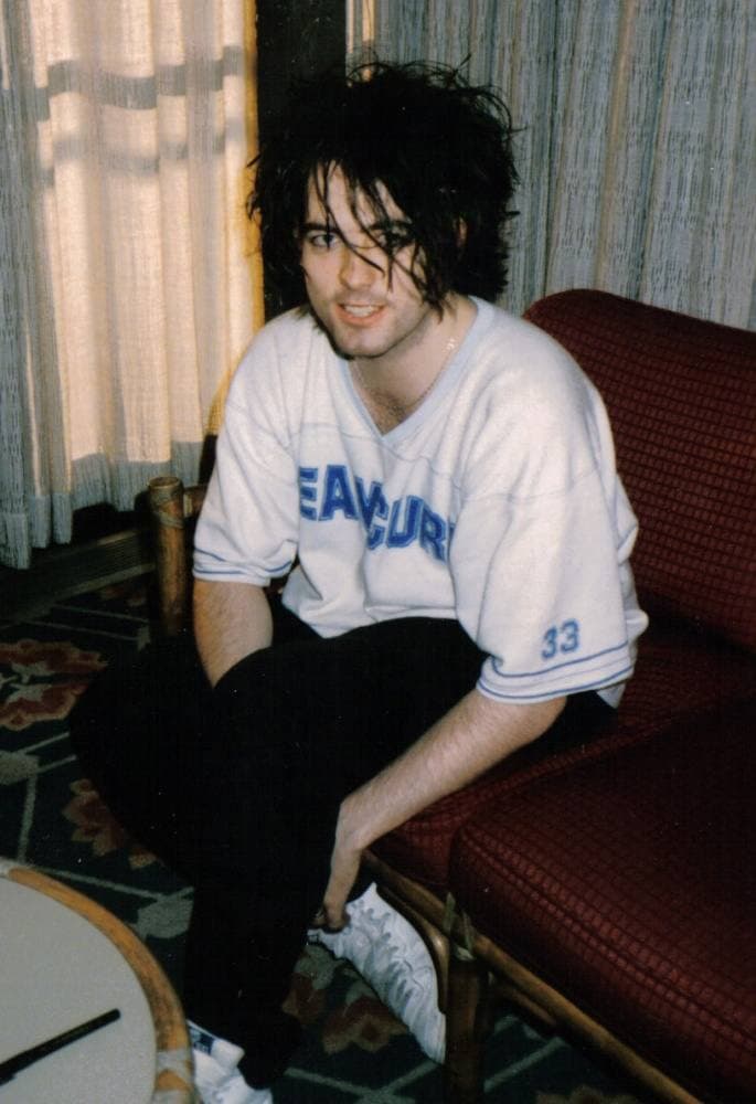 Robert Smith in 1985, his black hair a wild tangle over his eyes, smiling in a white football shirt with blue lettering, sitting on a red hotel chair