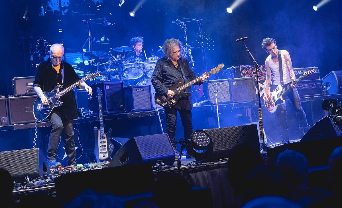 The Cure on a blue-lit stage, Robert Smith in the centre with a guitar between two guitarists, the drum kit behind them