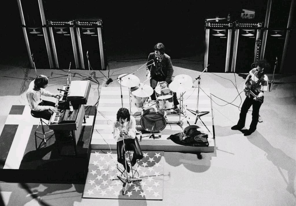 The Doors on a bare white television studio floor from above: Manzarek at the organ, Densmore at the drums on a flag rug, Krieger with his guitar and Morrison at the microphone