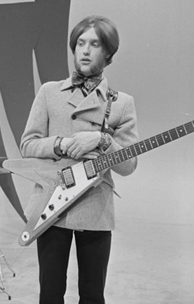 Dave Davies with long fringe and neckerchief, holding a Flying V guitar in a studio