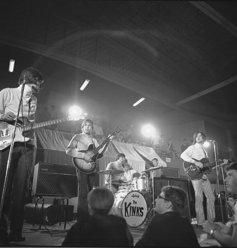 The Kinks playing on a hall stage under lights, with a drum kit marked The Kinks, black and white