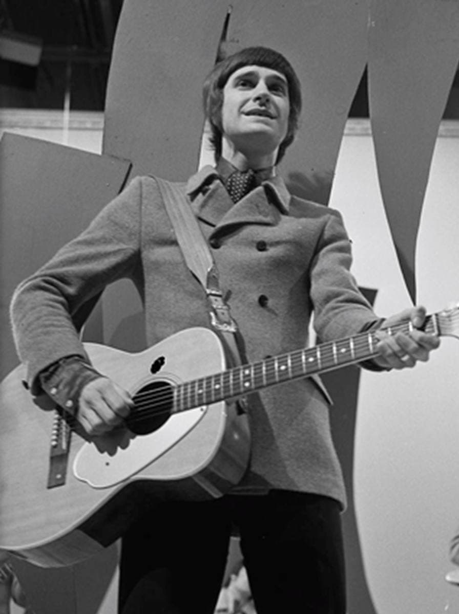 Ray Davies in a double-breasted coat and cravat, singing with an acoustic guitar in a television studio