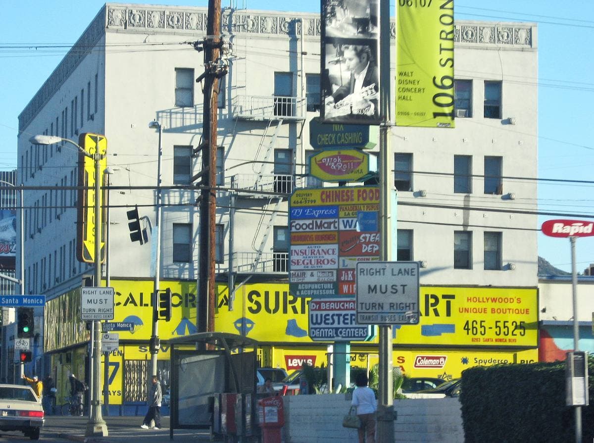 A busy Hollywood street corner with a pale office building, a strip-mall sign and traffic lights