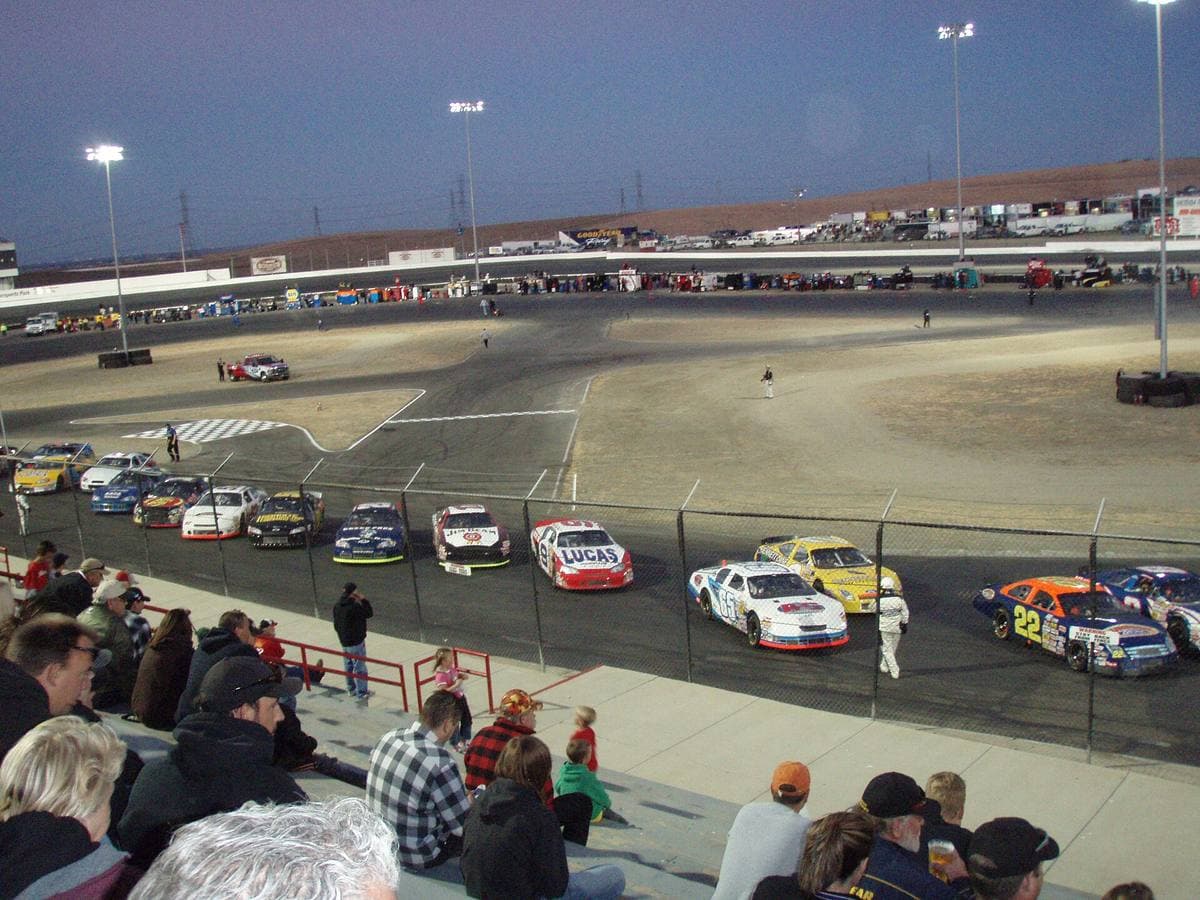 Stock cars lined up on an oval track under floodlights, with a small crowd in the stand