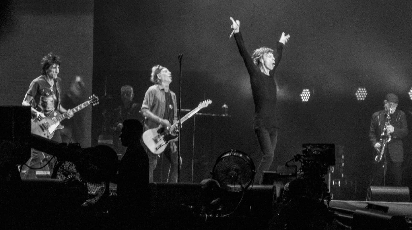 The band on a dark stage, Mick Jagger with both arms raised