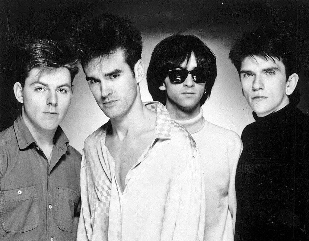 The four Smiths shoulder to shoulder against a pale wall, Marr in round dark glasses, Morrissey in an open white shirt