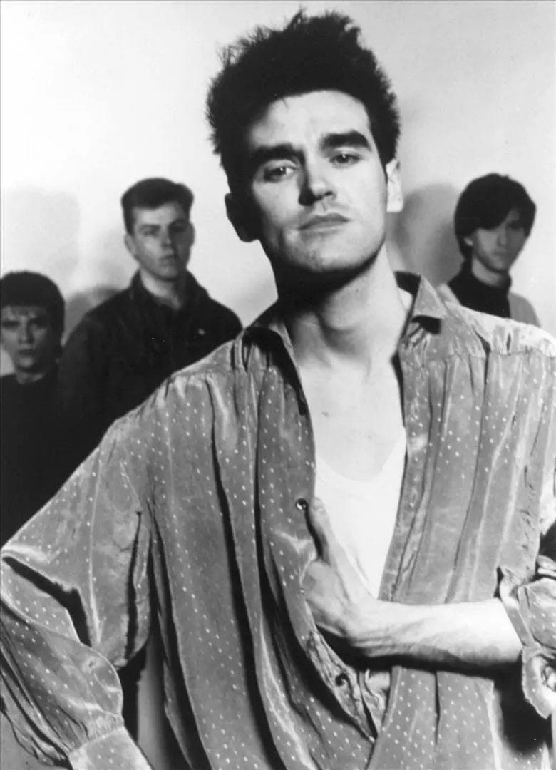 Morrissey in a loose silky shirt, hand on his chest, with Joyce, Rourke and Marr standing in shadow behind him