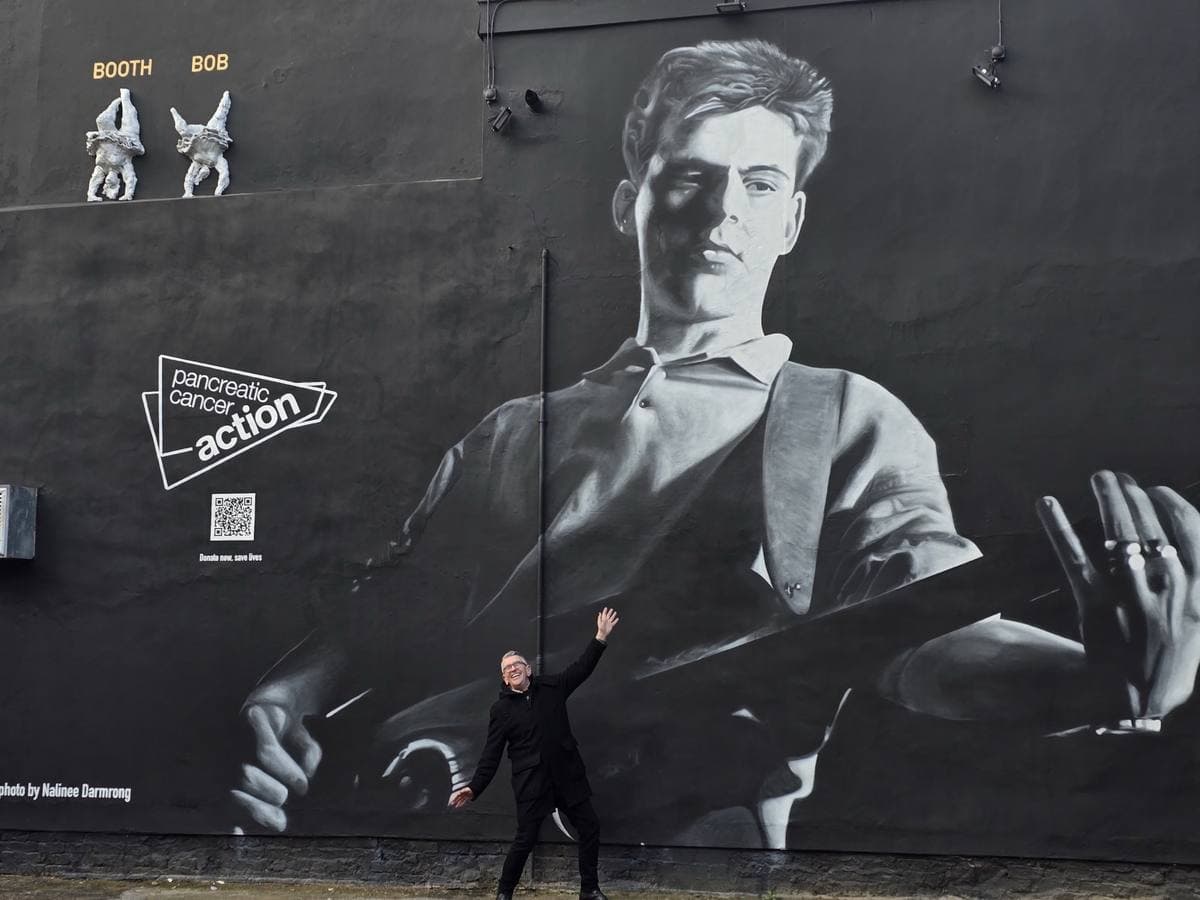 Mike Joyce in a black coat, arm raised, in front of the giant mural of Andy Rourke