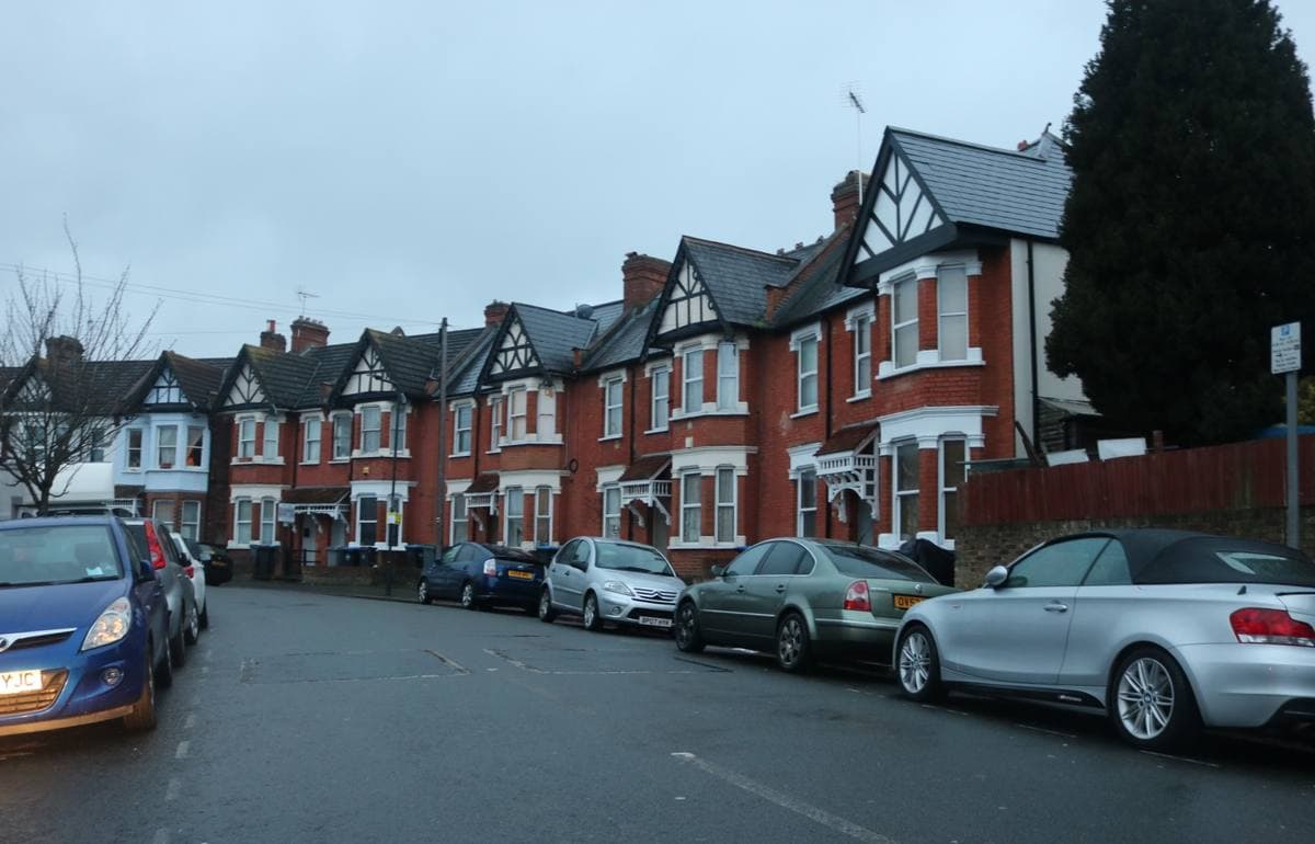 A residential street of red-brick houses with parked cars under a grey sky