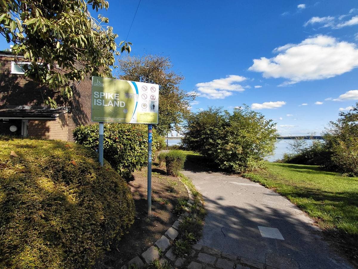 A path between bushes past a green Spike Island sign, the river glinting behind
