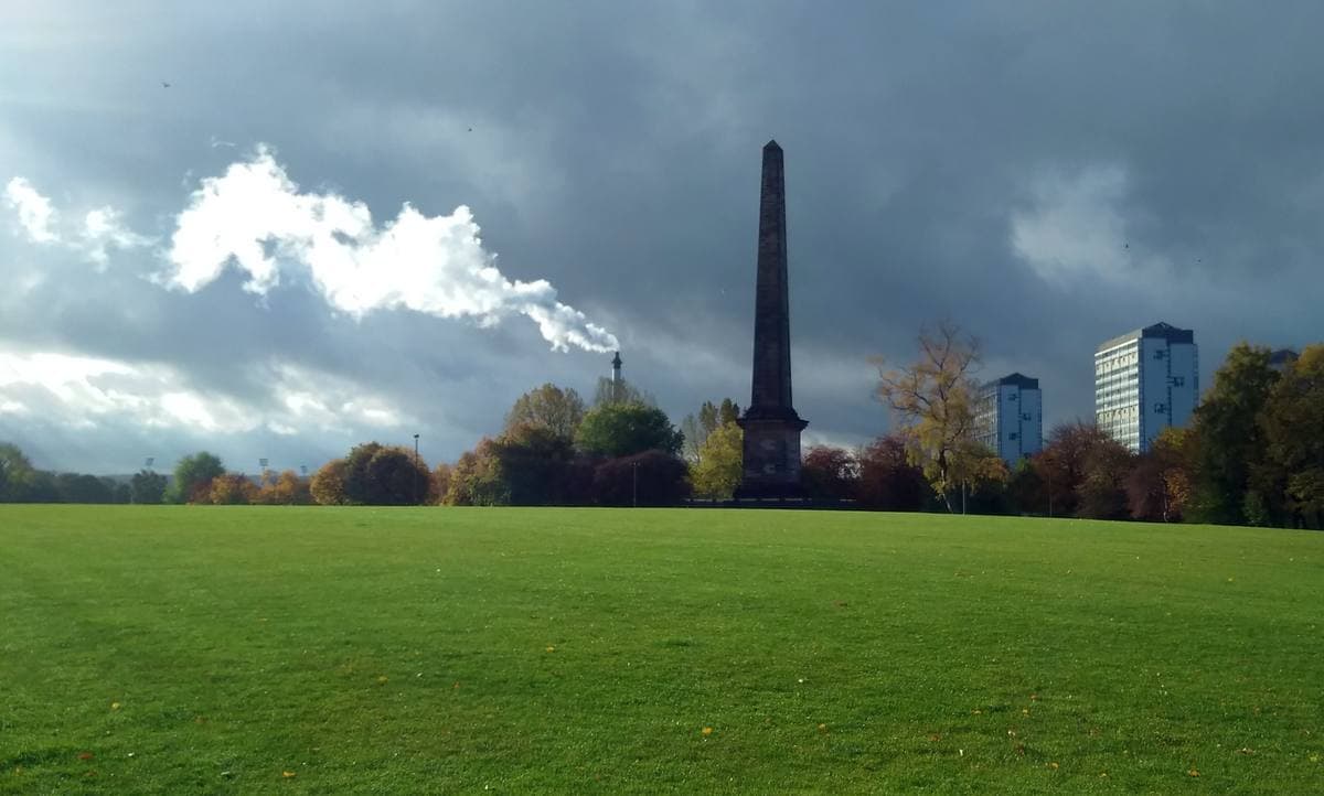 A broad green lawn under heavy cloud, a tall stone obelisk among autumn trees
