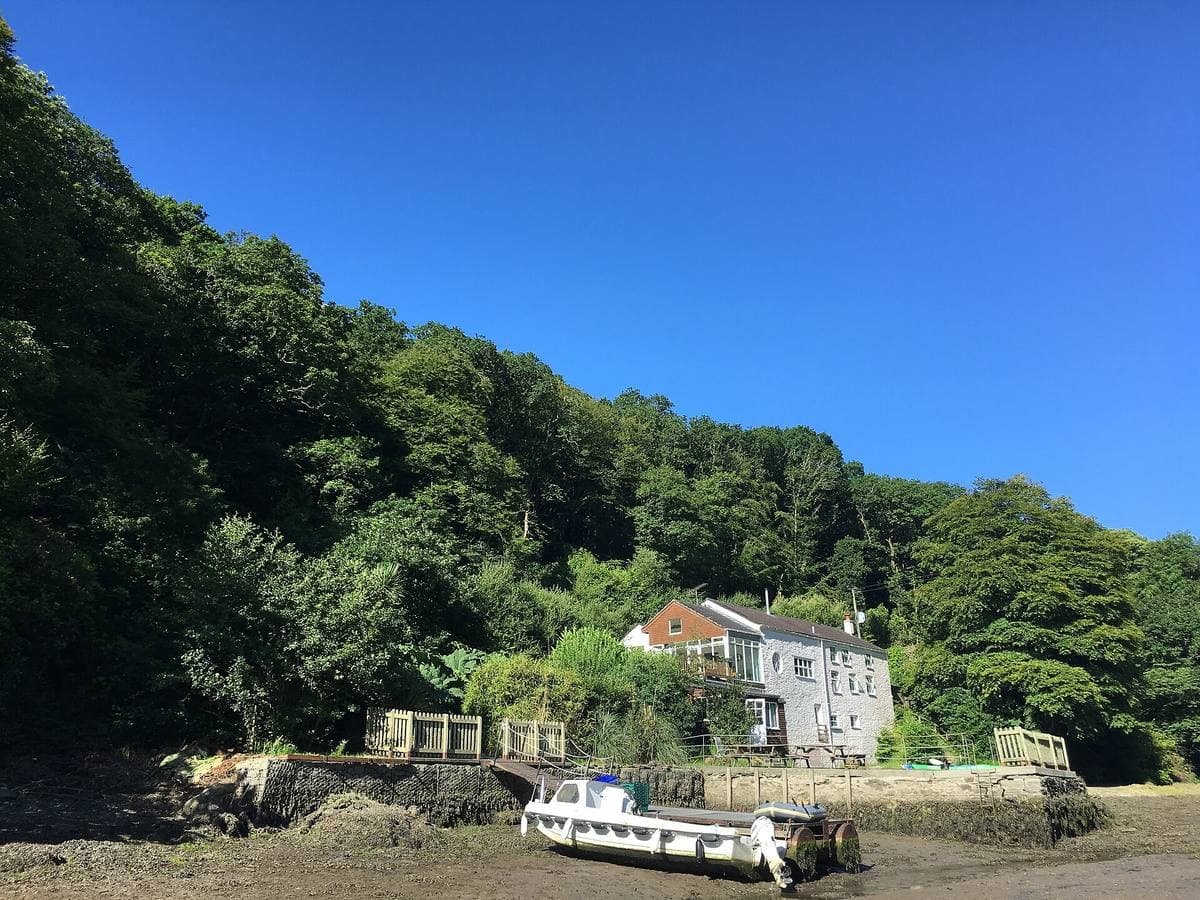 A white house among dense woods at the edge of a tidal creek, a small motor boat on the mud