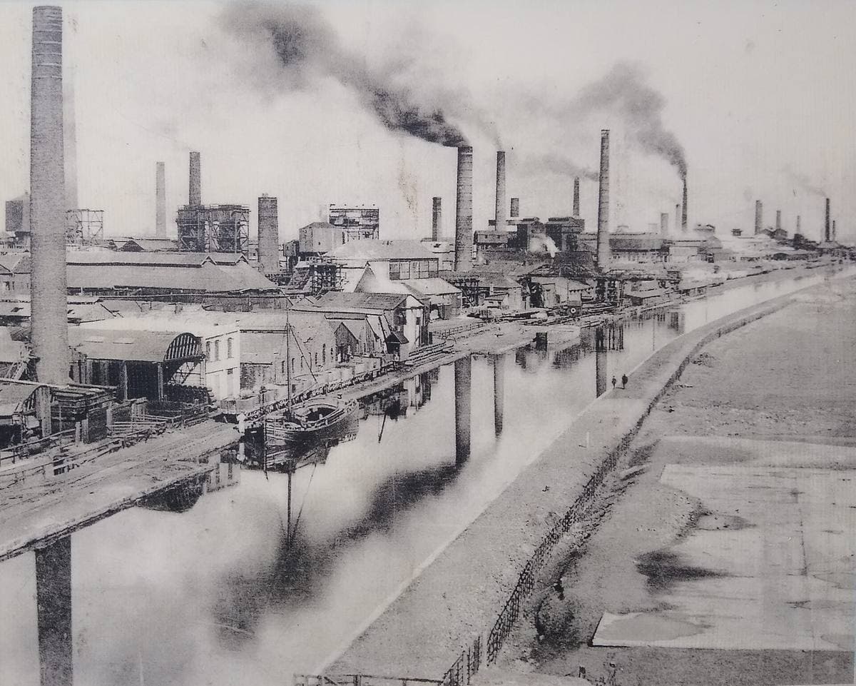 An old black and white photograph of tall smoking chimneys and works buildings beside a straight canal