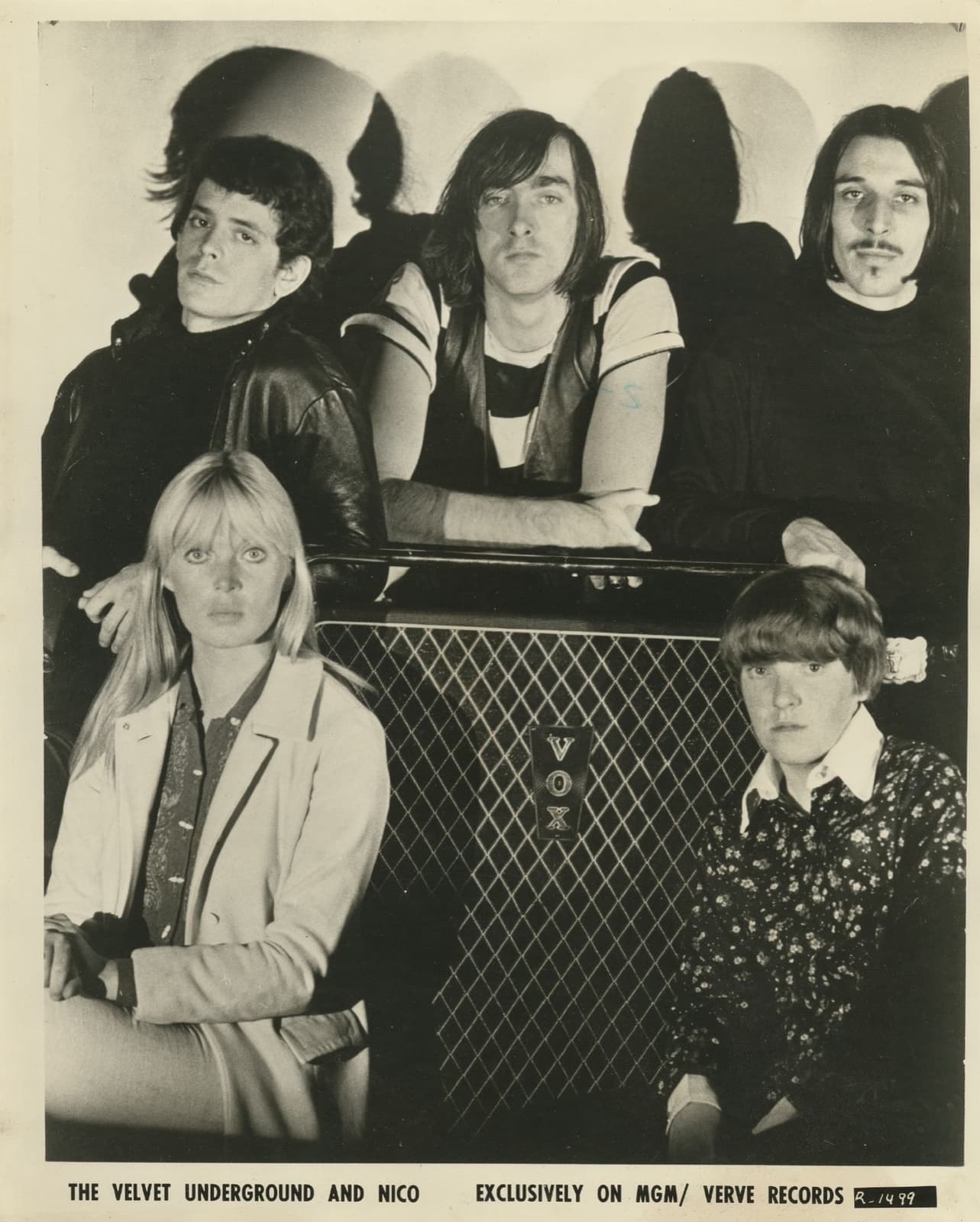Five young musicians in black and white: three men stand behind a speaker cabinet, a blonde woman and a short-haired drummer sit in front