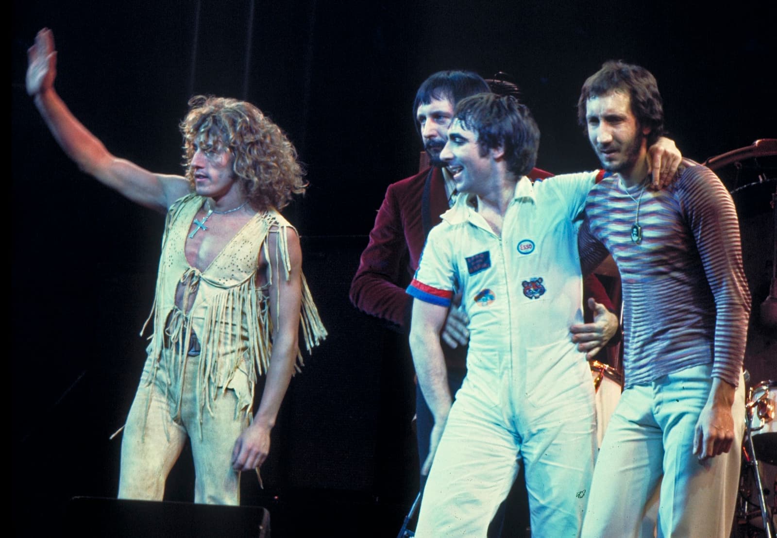 Roger Daltrey in a fringed suit waving, beside John Entwistle, Keith Moon in a white boiler suit and Pete Townshend in a striped top, arms round each other on stage