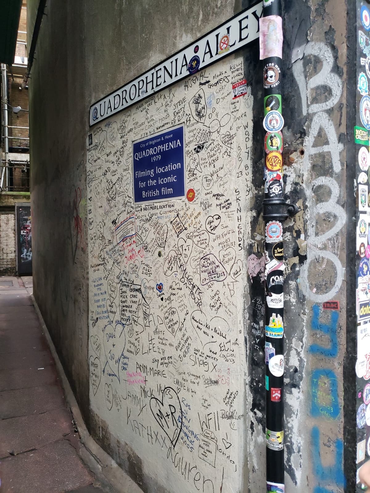 A narrow alley wall covered in names and graffiti under a Quadrophenia Alley sign
