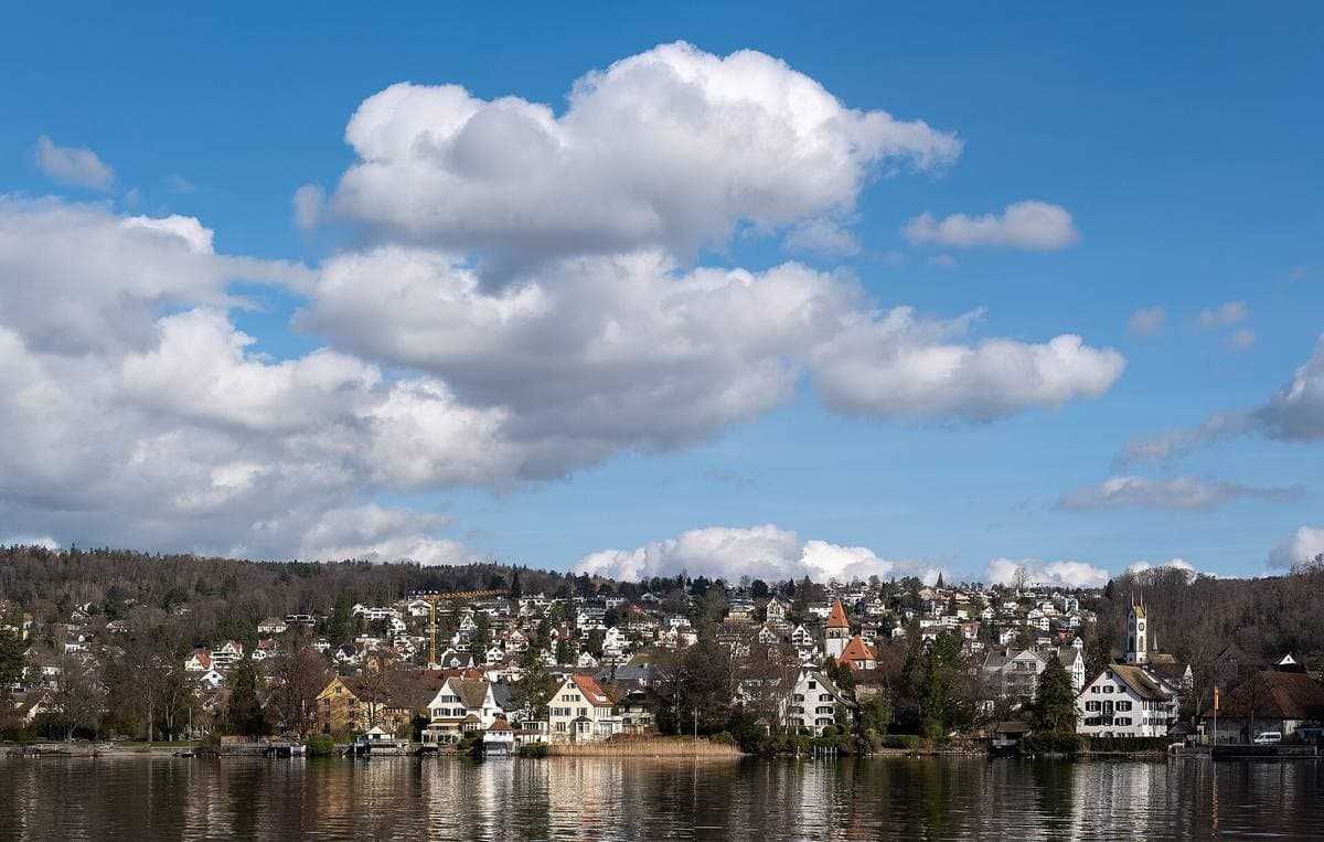 A lakeside village of white houses below wooded hills, seen from the water