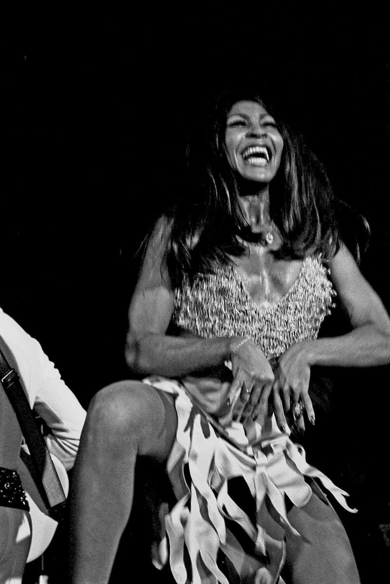 Tina Turner laughing on stage in a short fringed dress, in black and white