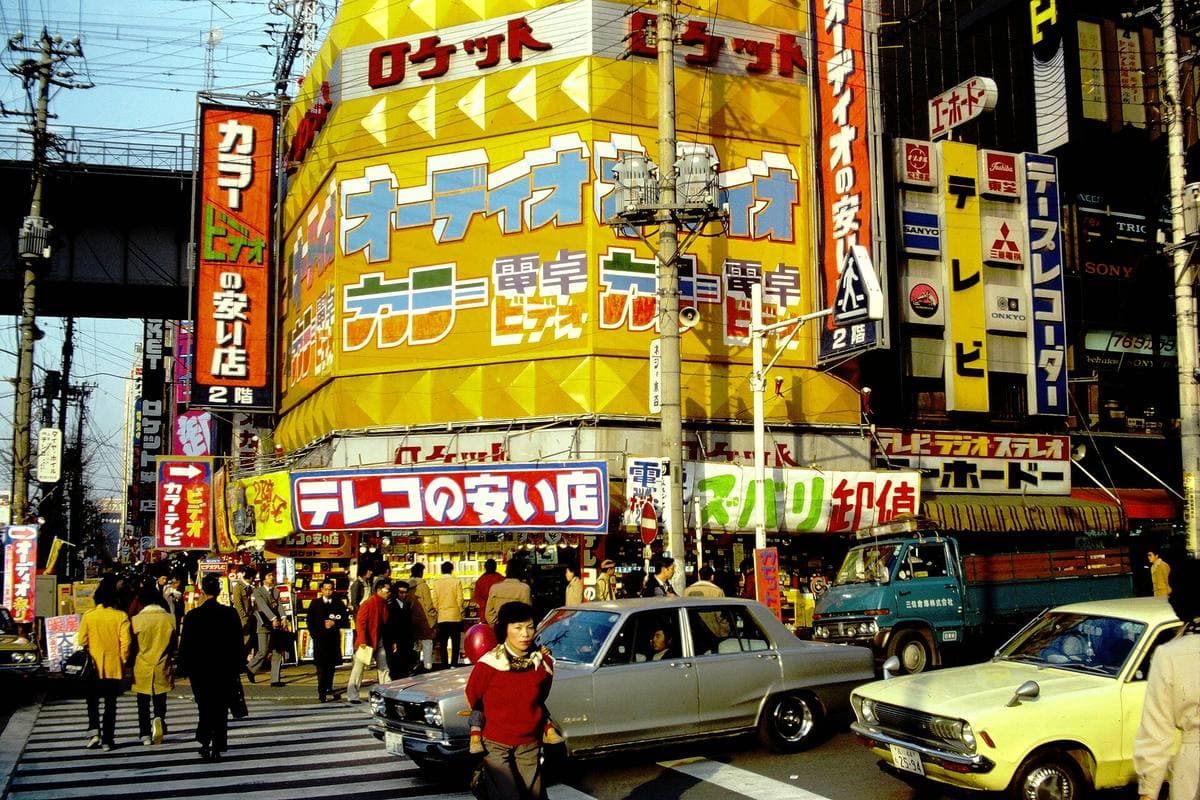 Crowds and cars below tall yellow and red shop signs in Tokyo's electronics district