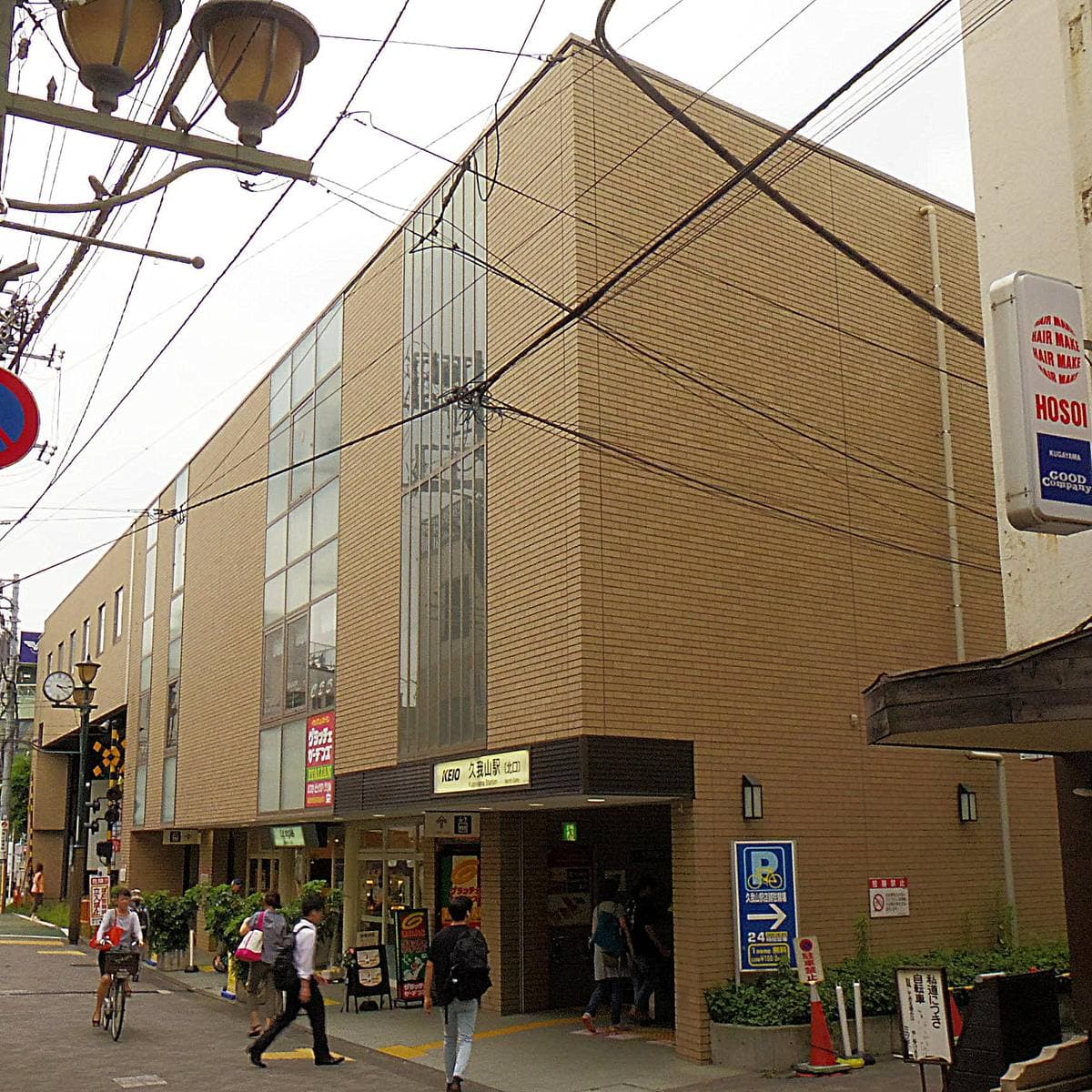 A low tan station building with people and bicycles passing on a sunny street