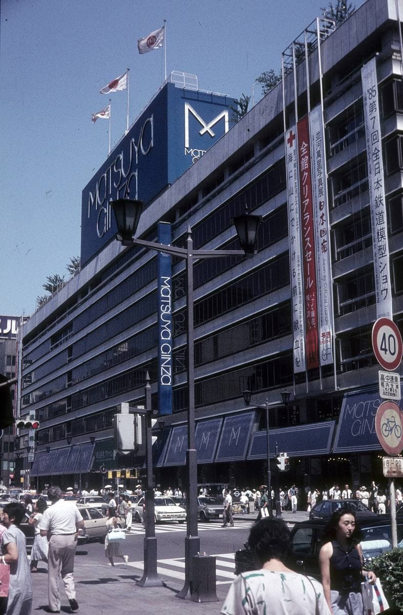 A department store frontage hung with vertical banners above a crowded Ginza pavement in summer