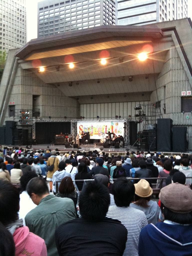 An outdoor concert shell at dusk with a crowd in front and a band far off on the lit stage